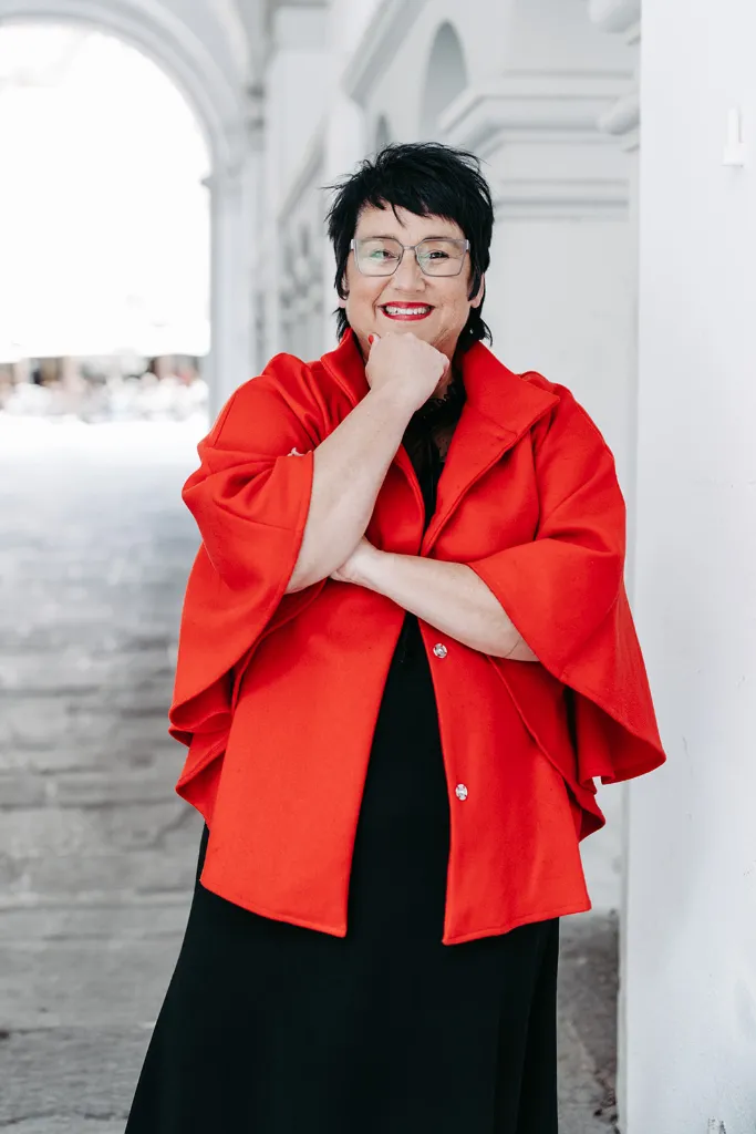 Sprecherin Sabine Jung-Bass schaut lächelnd direkt ins Bild. Sie trägt einen roten Mantel